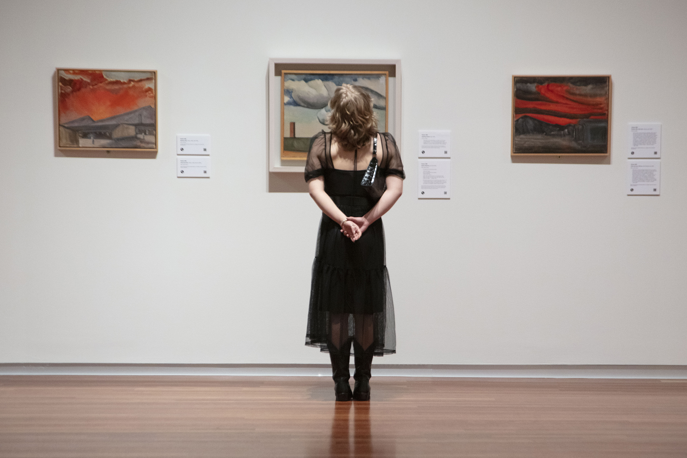 A photo of a white woman in a black dress with light blonde hair standing with her back to the camera, facing three framed paintings hanging on a wall.