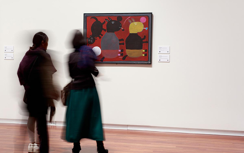 An image of two blurry figures in an open gallery, facing a large, framed painting of animal-like figures in bold colors.