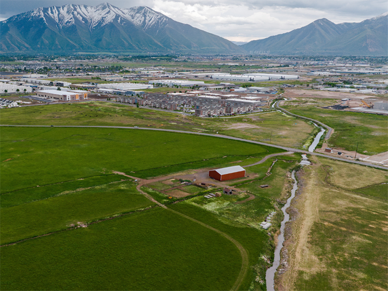 Aerial view of a green field with a red barn in the middle and mountains and city in the background.