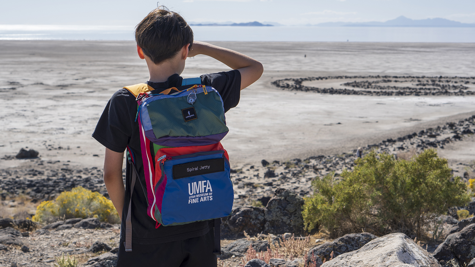 a boy about 10 years old stands on a hill overlooking Great Salt Lake and the Spiral Jetty he is wearing a bright multi backpack