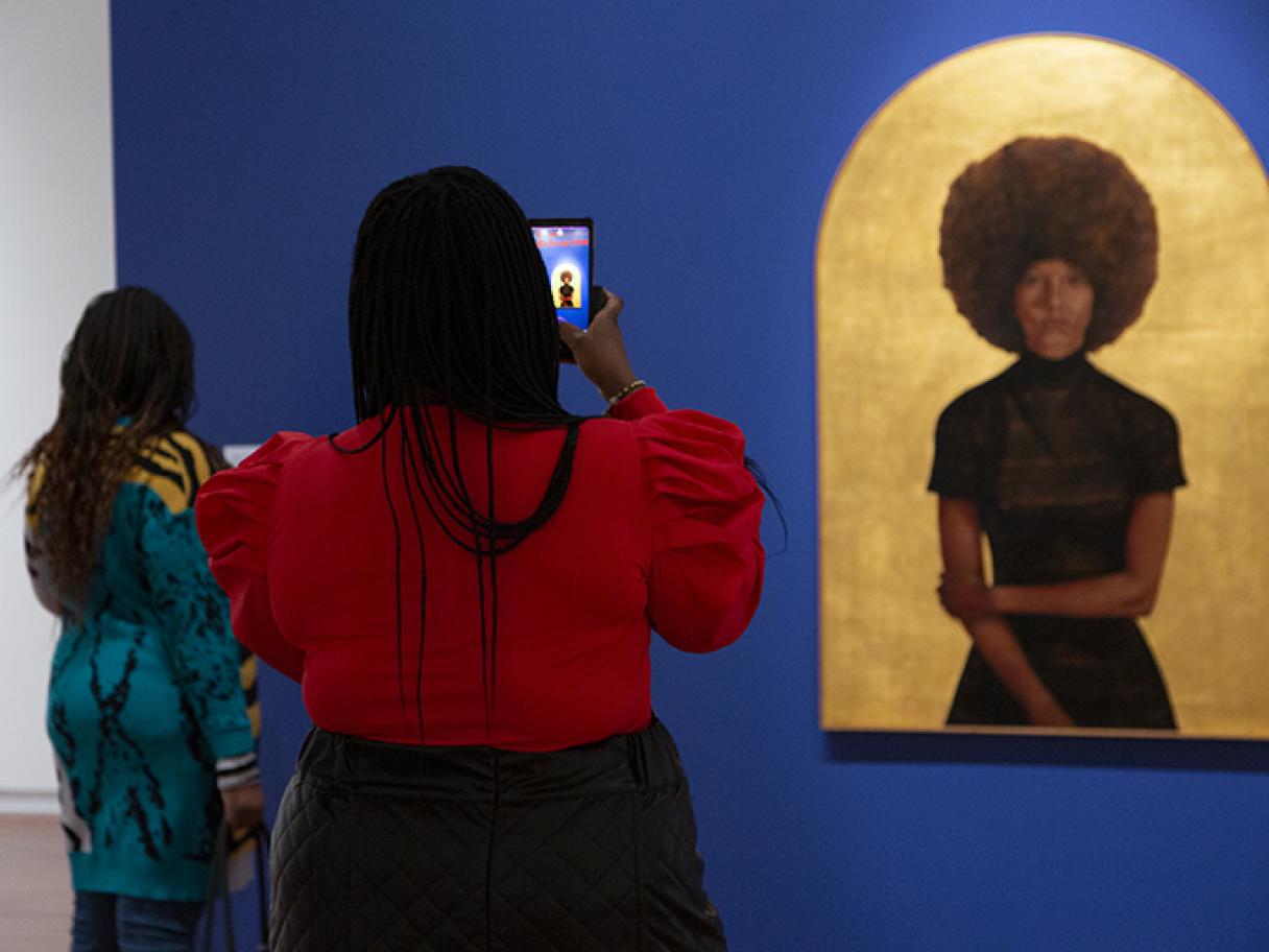 A woman in a red shirt holding up a phone to video Lawdy Mama, a portrait of a black woman with an afro on a gold leaf background. 