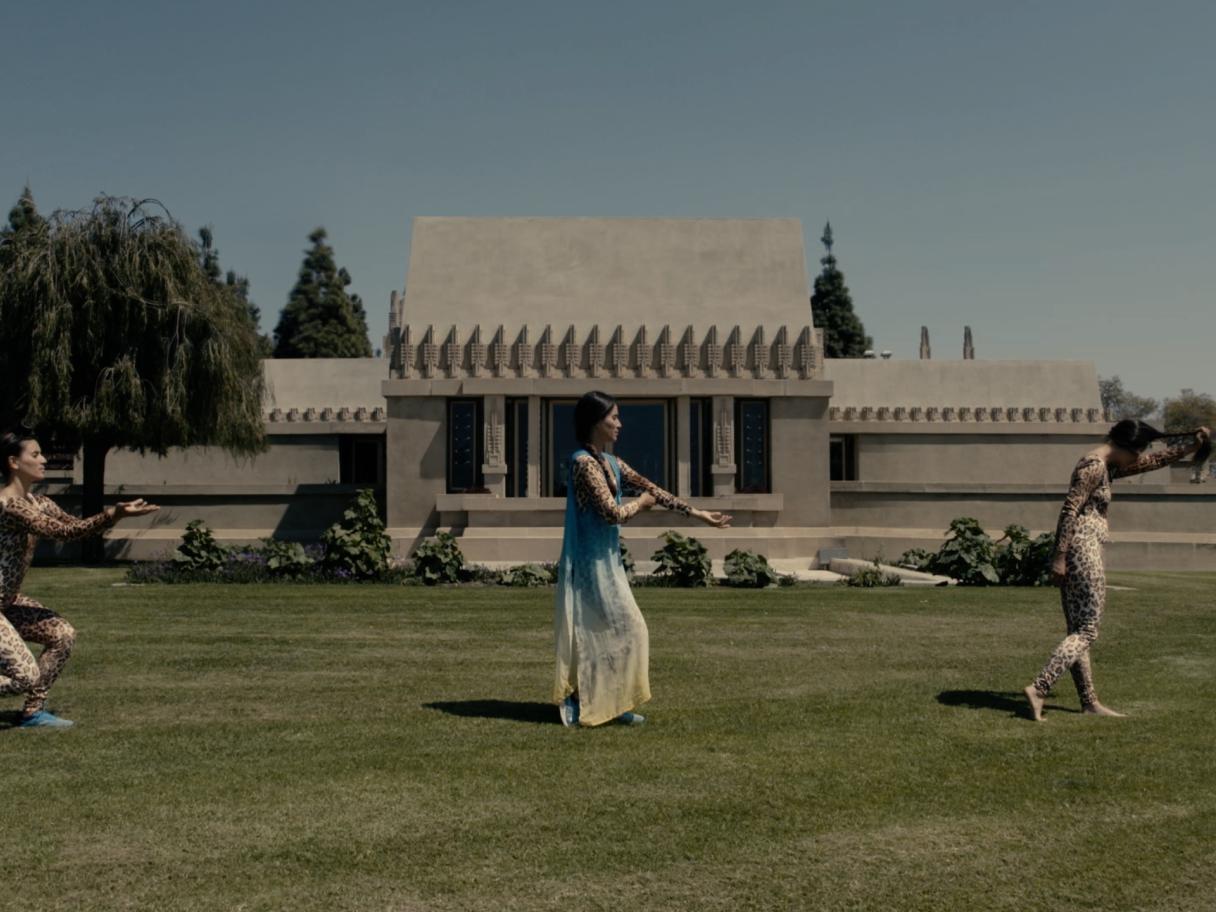 Three women dancing through a grass lawn in front fo a gray, cement building