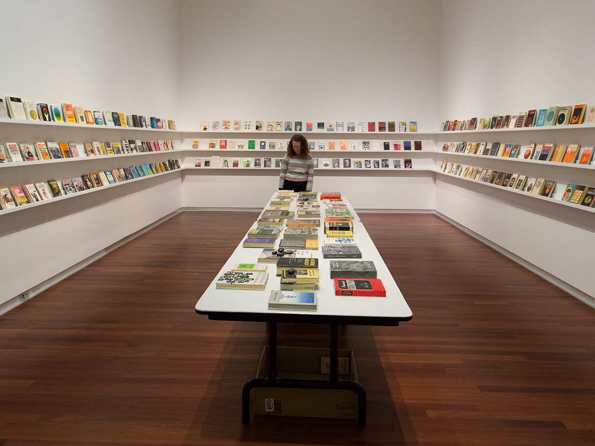 A person stands at the end of a long white table covered in books. The surrounding three walls have three continuous white shelves filled with colorful books too