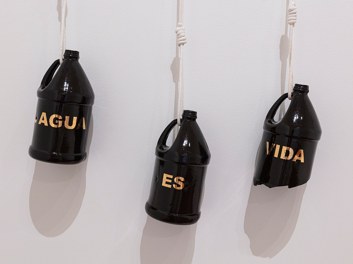 Three dark bottles with gold letters on them hang from white rope on a white wall. One of the bottles is broken.
