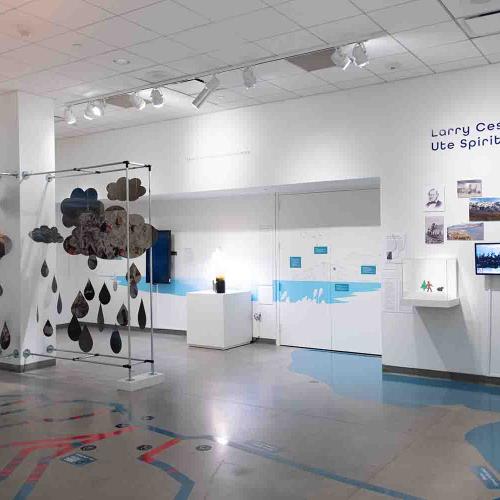 Confluence gallery, white room with blue vinyl map of rivers and a lake in the floor a metal cloud sculpture on the left with a wall of landscape images on the right