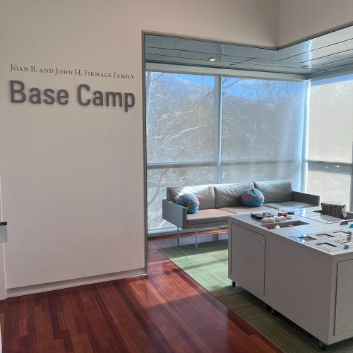 A photo of a room with white walls, wood floors and floor to ceiling windows on two walls. The word Base Camp is printed on the wall in grey letters. There's a light grey couch on the left and a grey table with different items in front of it. 