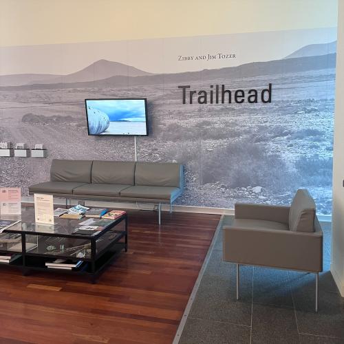 A photo of a room with a grey couch against the far wall and grey arm chairs on either side of a glass coffee table with books on top. The word Trailhead is printed in dark letters on the far wall over a mural of a desert landscape with mountains.